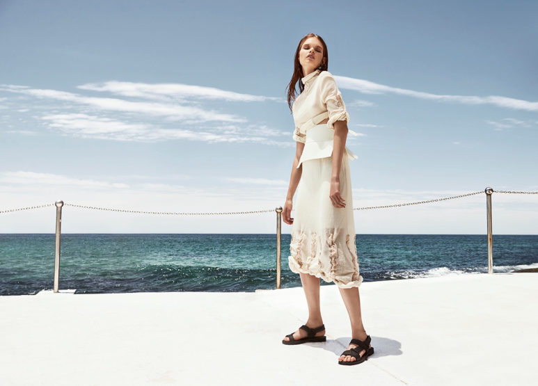 A fashionable adult female poses on a white concrete structure with ocean and a cloudy sky in the background, wearing a white outfit.