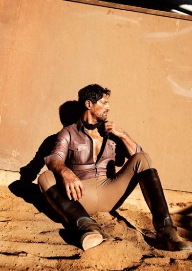 Portrait of an adult male in a light purple button-down shirt and tan pants, sitting in the dirt and wearing tall black riding boots.