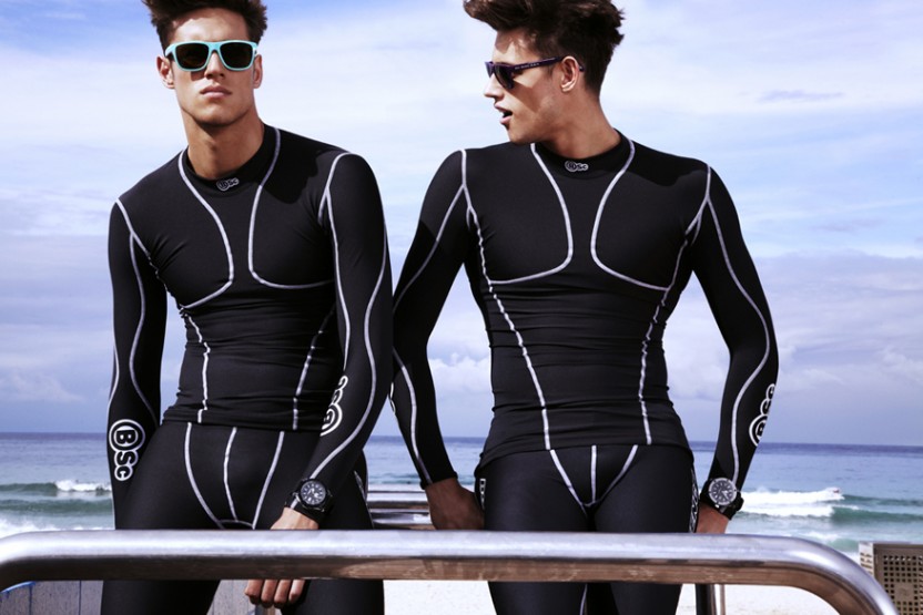 Two adult male models pose at the beach wearing black and white athletic compression shirts and pants with sunglasses on a sunny day.