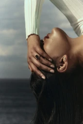 Close-up profile of a woman with dark hair touching her face, wearing a ring and a ribbed white sleeve against an ocean horizon