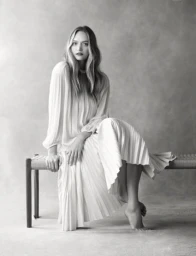 Black and white portrait of a woman in a long pleated dress seated on a wooden woven bench against a textured backdrop.