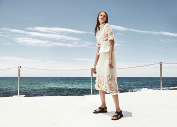 Model in cream dress and black sandals standing on a coastal platform against the ocean and sky