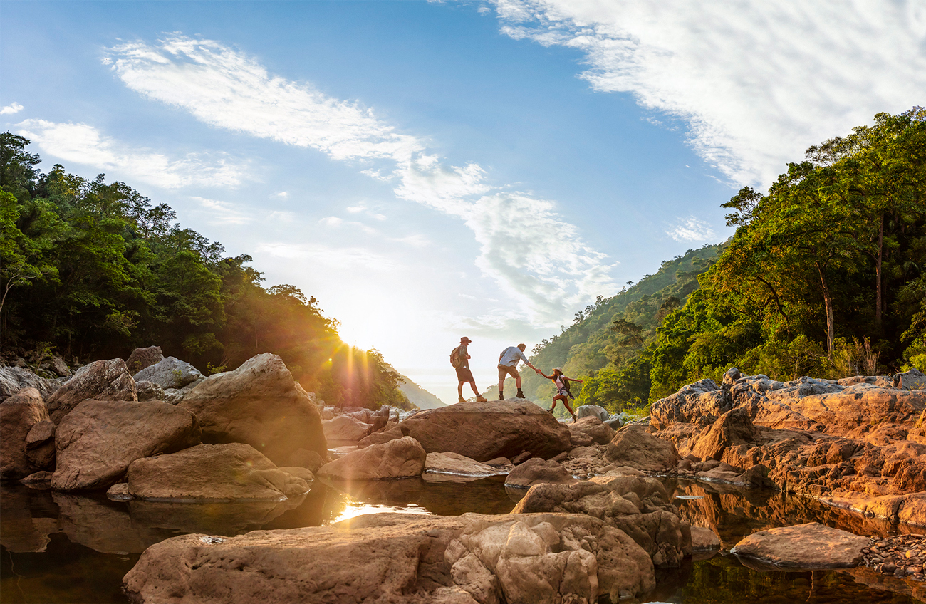 Three people hiking in a mountainous region, crossing rocks in a shallow river with lush forest and clear blue sky in the background.