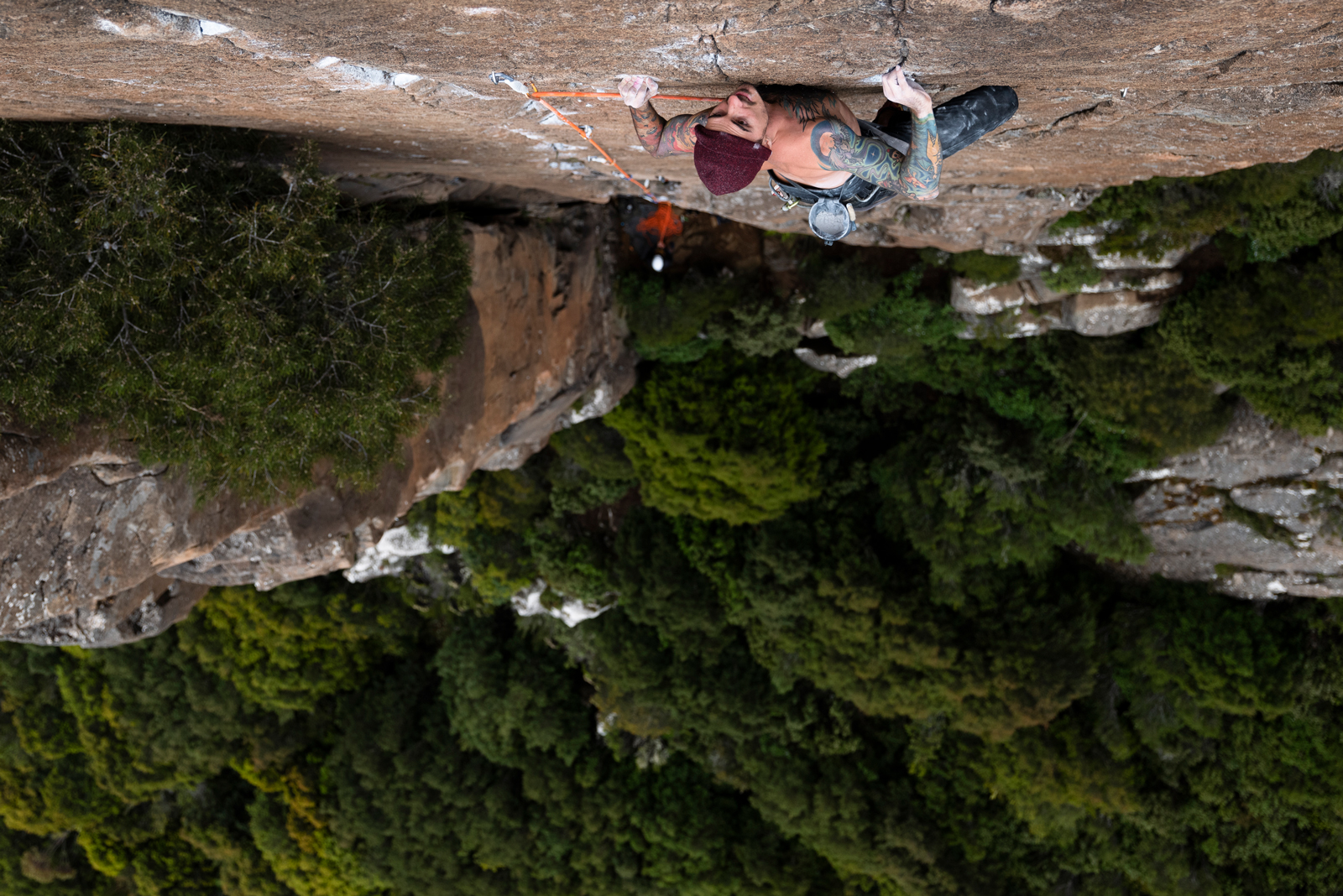 Adult male with tattoos rock climbing outdoors on a rock face with trees in the background wearing a burgundy knit hat.