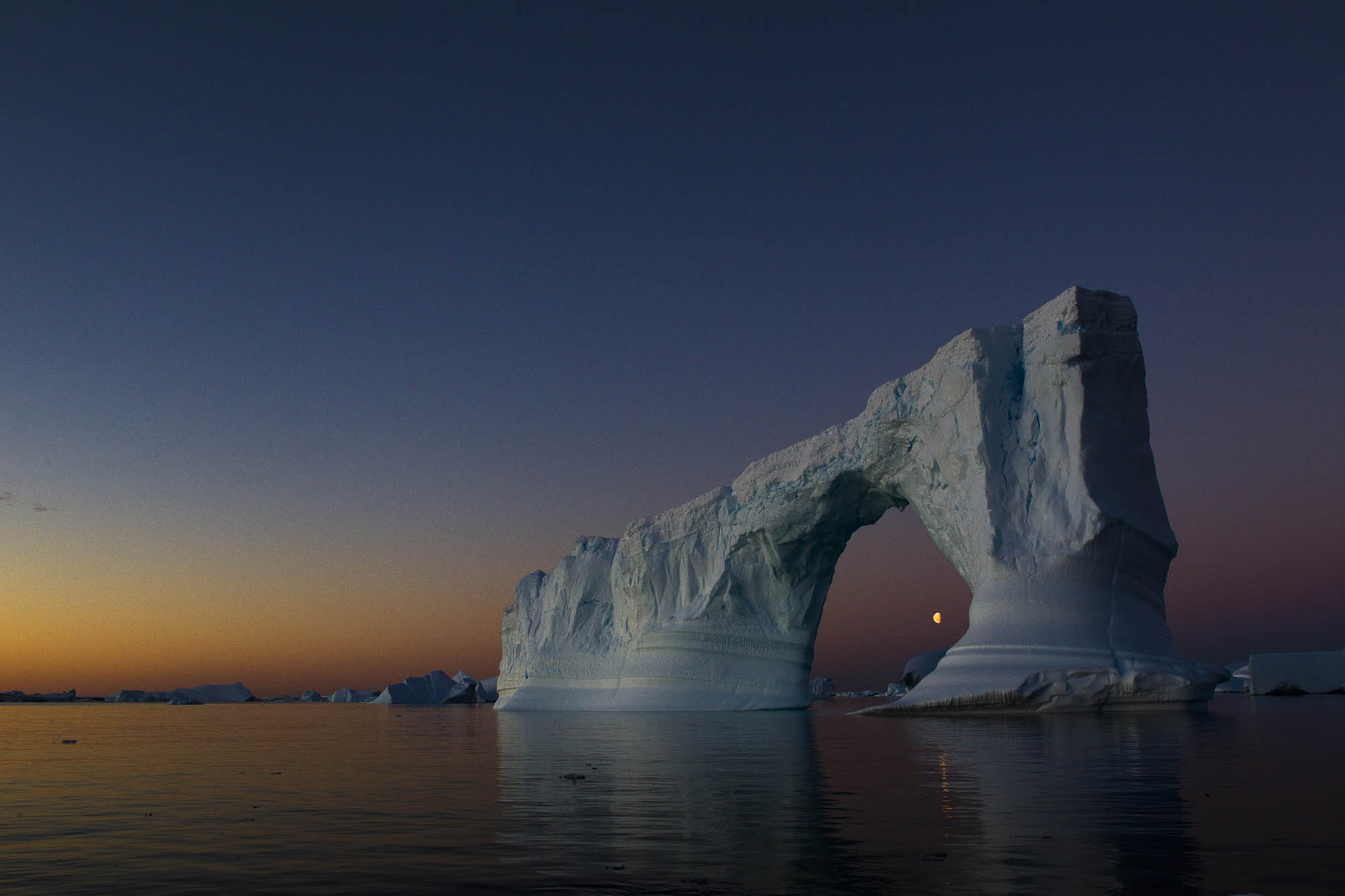 Dramatic iceberg archway formation against a vibrant twilight sky, reflecting in the calm waters of the polar region.