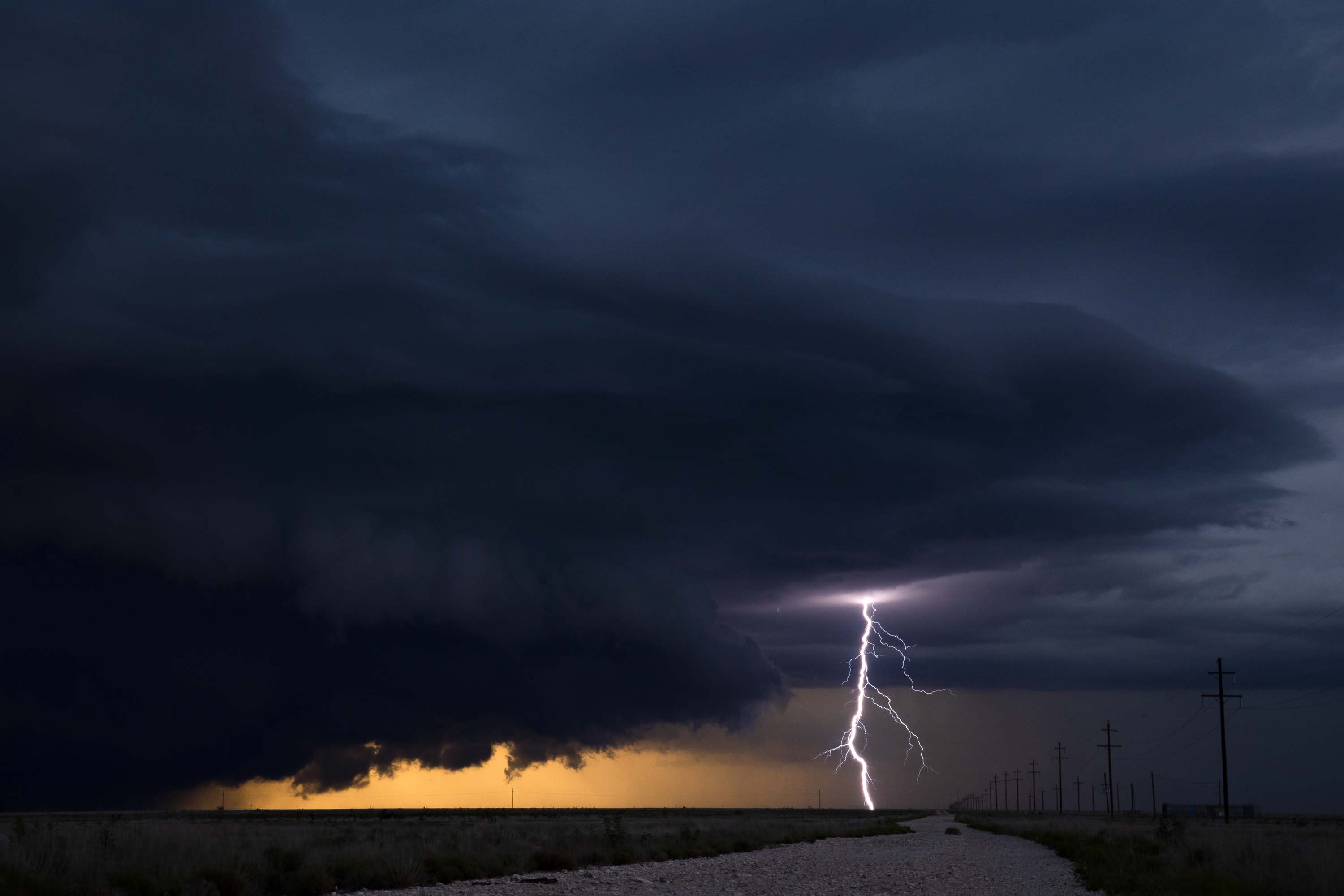 Dramatic storm scene: lightning strikes illuminate a rural road, dark storm clouds gather overhead, and a vibrant orange horizon glows.