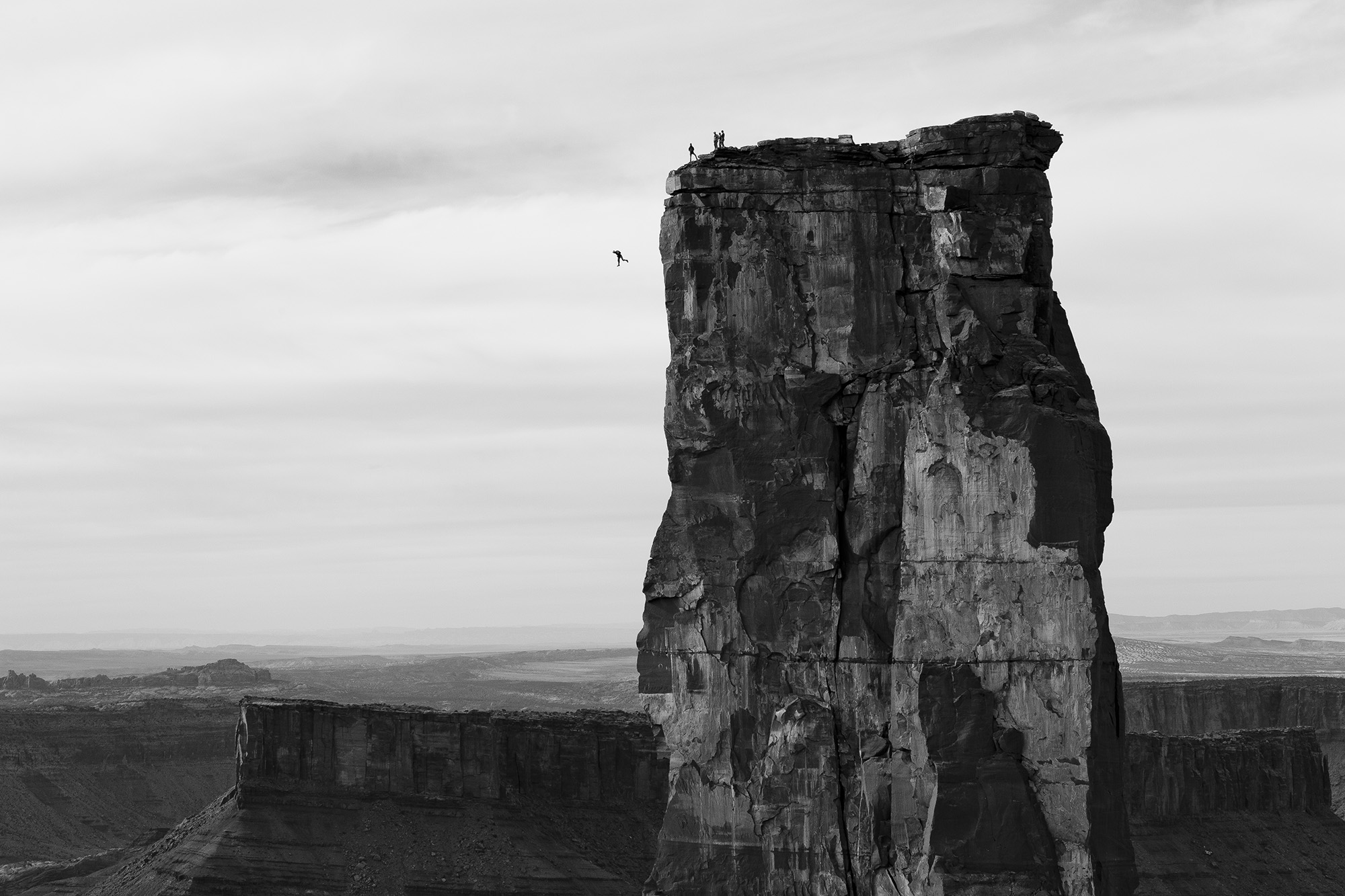 Monochrome landscape featuring a towering rock formation with figures at the summit and a base jumper in Utah, against a cloudy sky.