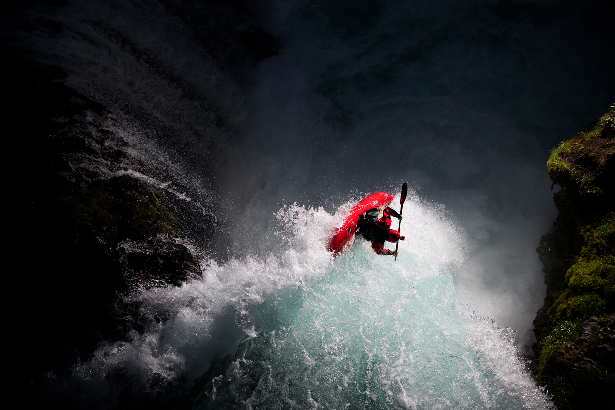 A kayaker in a red kayak plunges down a powerful waterfall, surrounded by white water and dark, rocky cliffs with green moss.