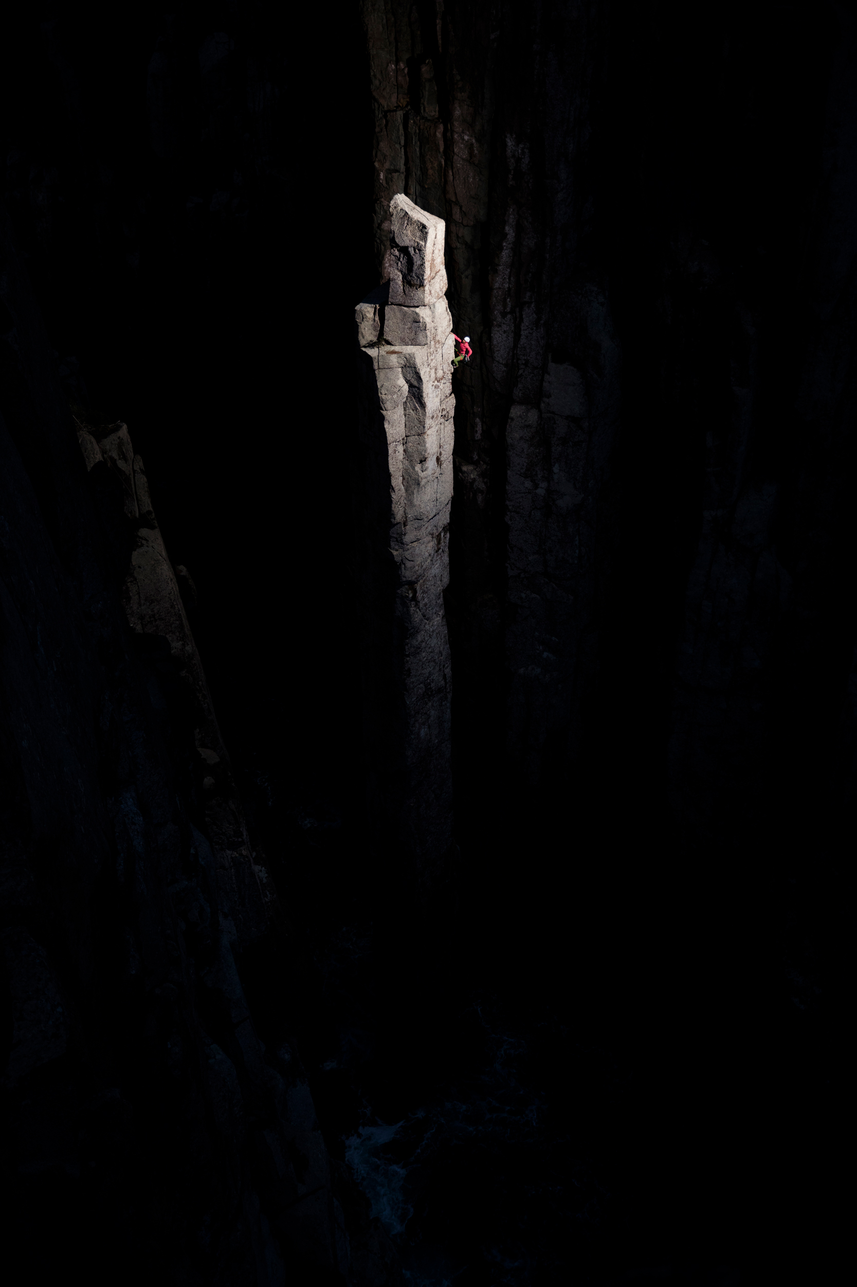 Rock climber ascends a tall, narrow rock formation bathed in light, surrounded by dark cliff walls in a dramatic outdoor setting.