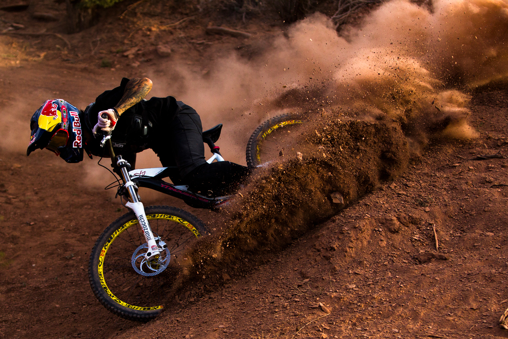 Mountain biker wearing a Red Bull helmet kicks up dirt while riding a white and yellow mountain bike on a dirt track outdoors.