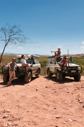Group of four people relaxing with drinks by two white off-road vehicles in an arid landscape with sparse trees and clear blue sky.
