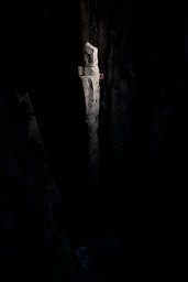 Rock climber in red apparel scaling a tall vertical stone pillar illuminated by a beam of light within a dark cavern.