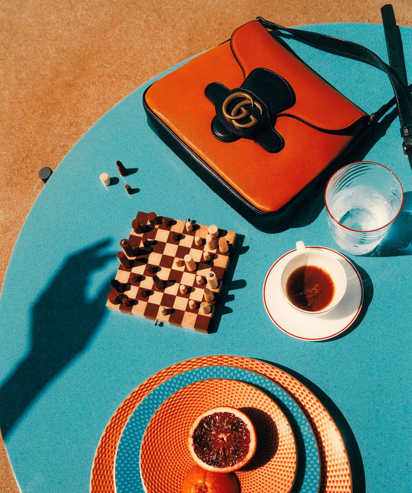 Overhead shot of an orange Gucci handbag, a chessboard, citrus fruit, a coffee cup, and a drinking glass on a blue surface.