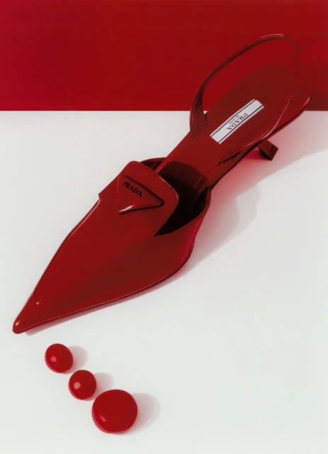 Close-up studio shot of a red Prada slingback heel with a pointed toe. Red decorative beads sit at the toe, against a white and red background.