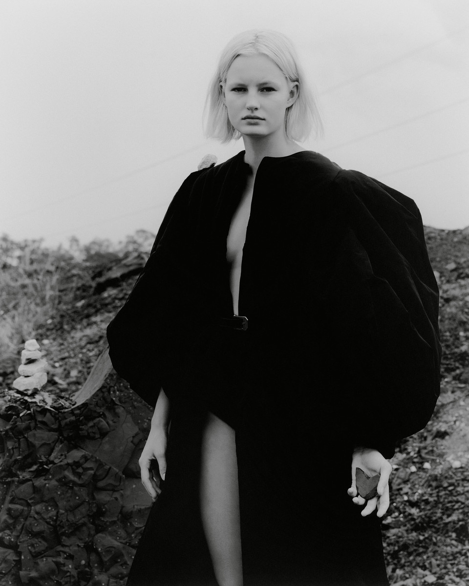 Monochrome outdoor portrait of a blonde woman in a long black robe, standing on rocky terrain under an overcast sky in a fashion shot.