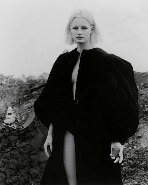 Monochrome outdoor portrait of a blonde woman in a long black robe, standing on rocky terrain under an overcast sky in a fashion shot.