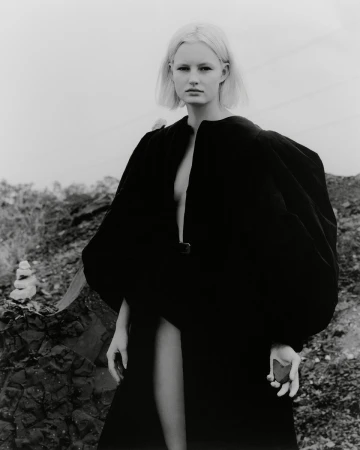 Monochrome outdoor portrait of a blonde woman in a long black robe, standing on rocky terrain under an overcast sky in a fashion shot.