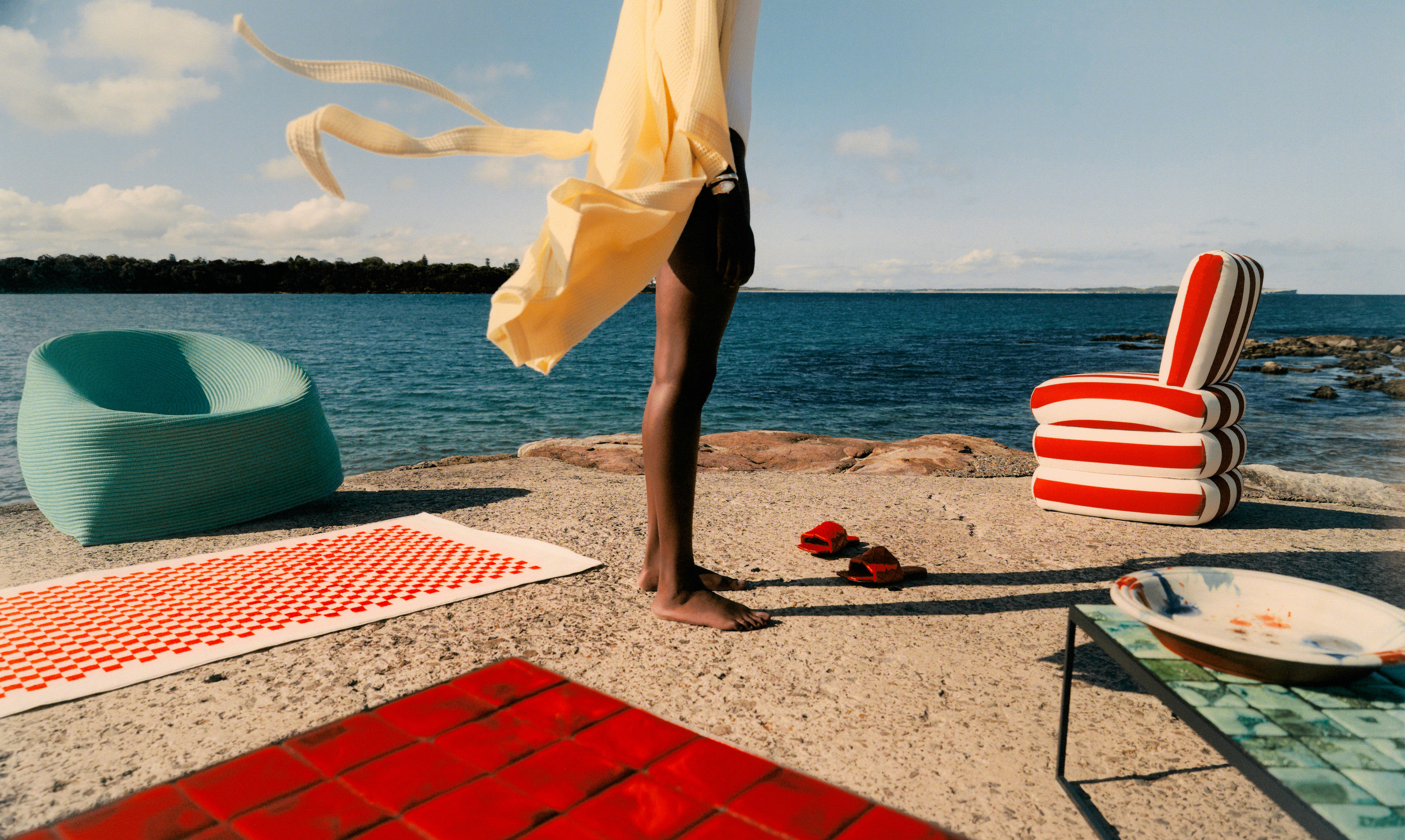 Outdoor scene featuring a person standing near the ocean with colorful chairs, a blanket, and a table on a sunny day.