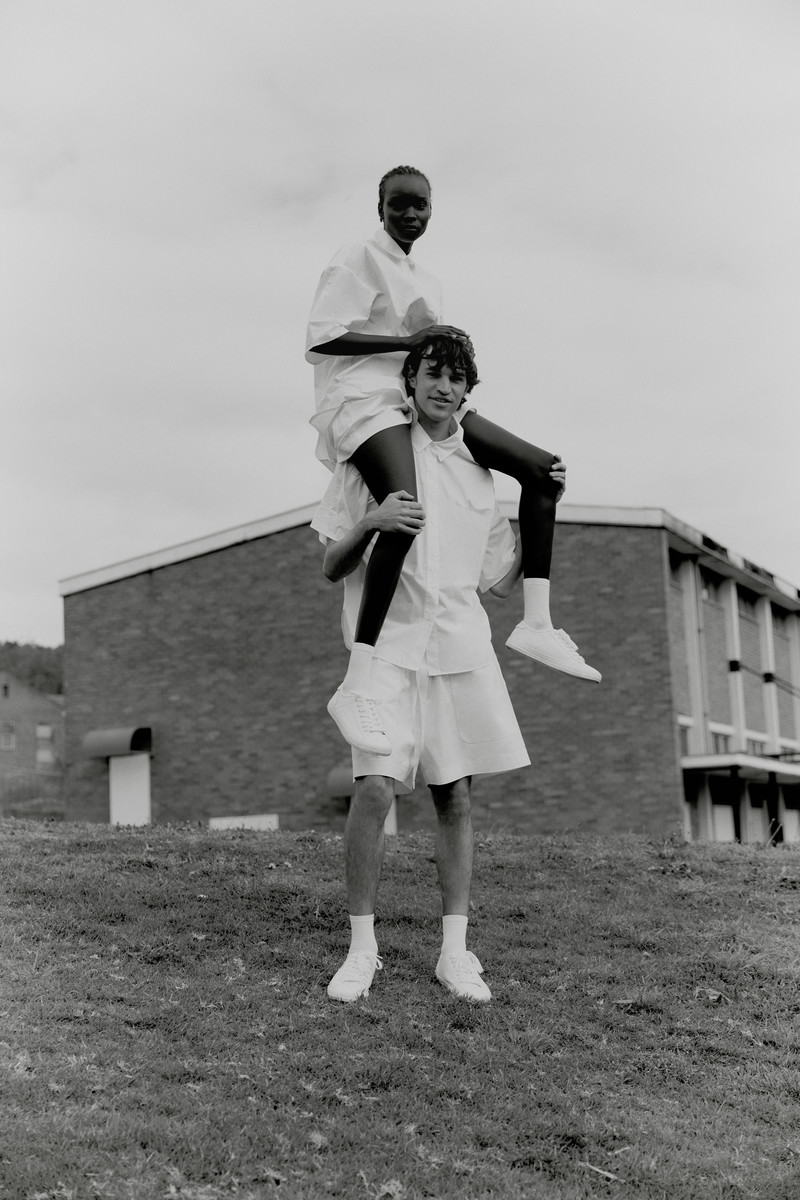 Two people outside, one woman sits on the shoulders of an adult male, both are wearing white shirts and shorts, with a building behind.