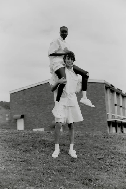 Two people outside, one woman sits on the shoulders of an adult male, both are wearing white shirts and shorts, with a building behind.