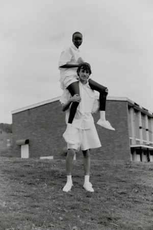 Two people outside, one woman sits on the shoulders of an adult male, both are wearing white shirts and shorts, with a building behind.