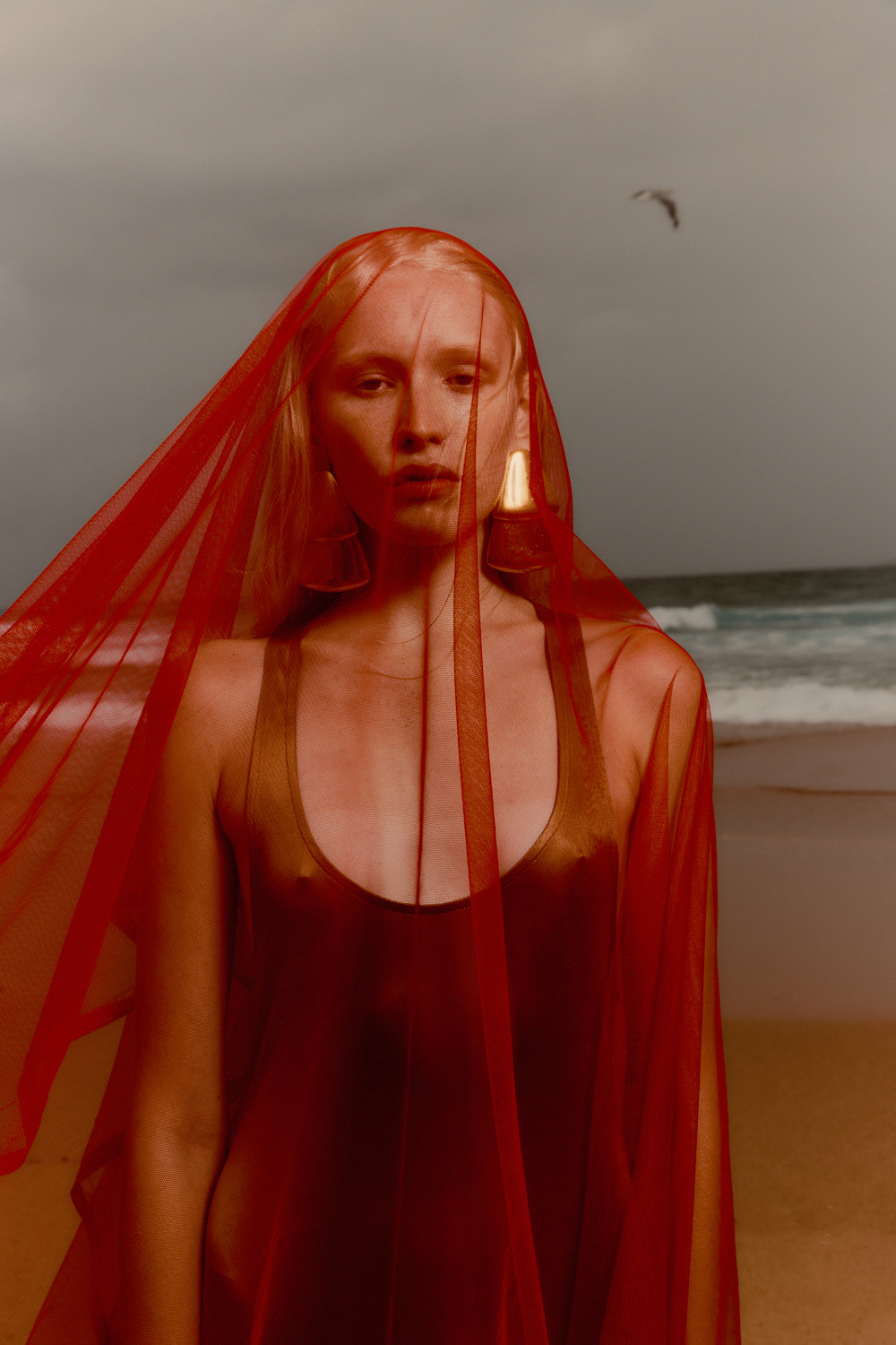 Woman with blonde hair wearing a red veil on a beach with the ocean and sky in the background. Beauty shot, fashion, lifestyle.