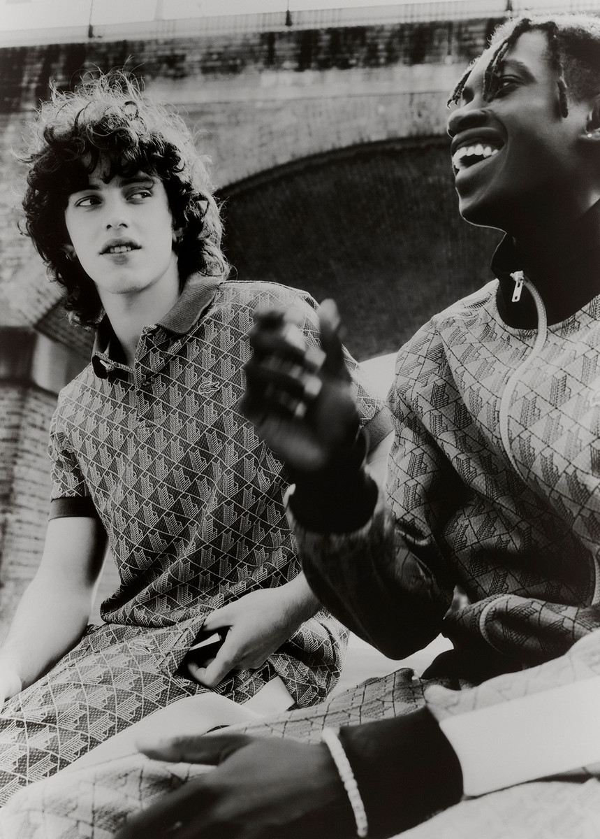 Two men wearing matching patterned outfits are sitting closely together with a building with an arch in the background in a monochrome photograph.