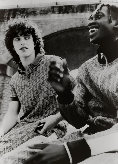 Two men wearing matching patterned outfits are sitting closely together with a building with an arch in the background in a monochrome photograph.
