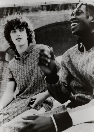 Two men wearing matching patterned outfits are sitting closely together with a building with an arch in the background in a monochrome photograph.