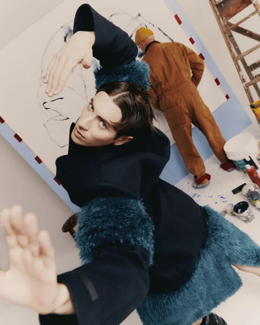Stylish man in blue furry coat poses with abstract art and painter at work in a brightly lit studio setting with ladder and paint cans.