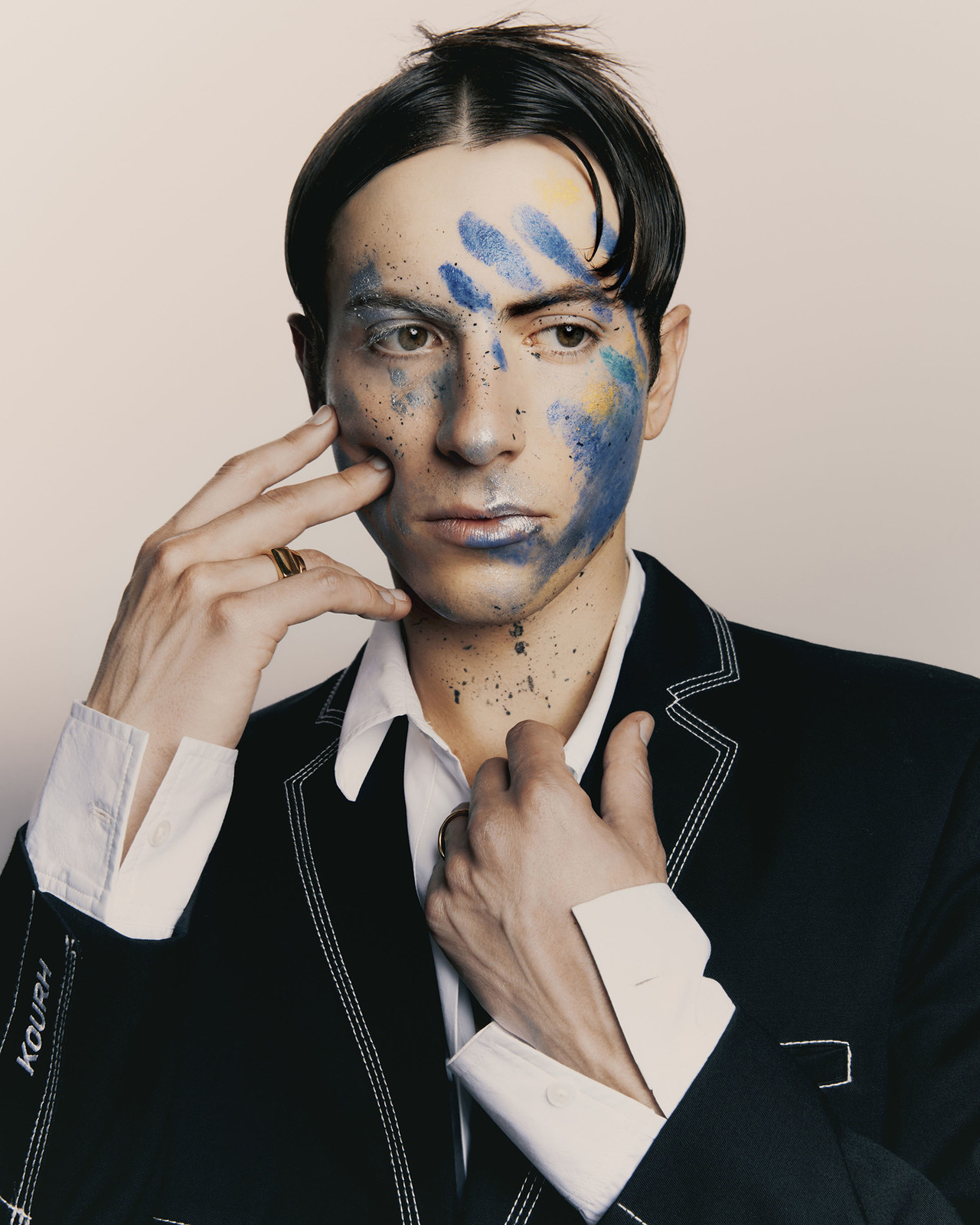 Portrait of a man wearing a dark jacket with white stitching, with blue face paint and a white shirt, photographed by Levon Baird