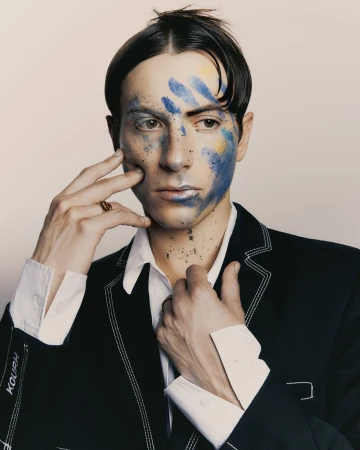 Portrait of a man wearing a dark jacket with white stitching, with blue face paint and a white shirt, photographed by Levon Baird
