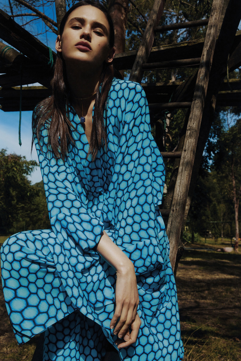 Young woman poses outdoors wearing a blue patterned dress and pendant necklace, treehouse in the background.