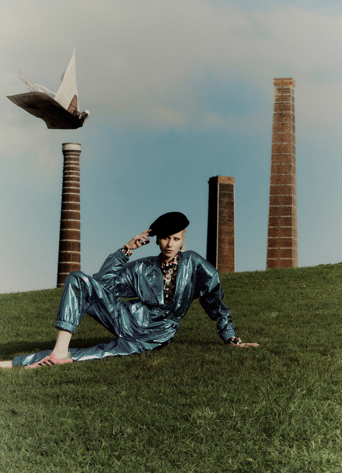 A woman in a blue jumpsuit poses on a grassy hill with industrial chimneys and a white bird-shaped sculpture in the background.