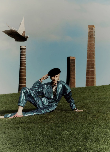 A woman in a blue jumpsuit poses on a grassy hill with industrial chimneys and a white bird-shaped sculpture in the background.