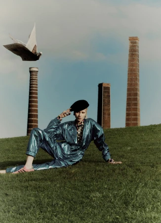 A woman in a blue jumpsuit poses on a grassy hill with industrial chimneys and a white bird-shaped sculpture in the background.