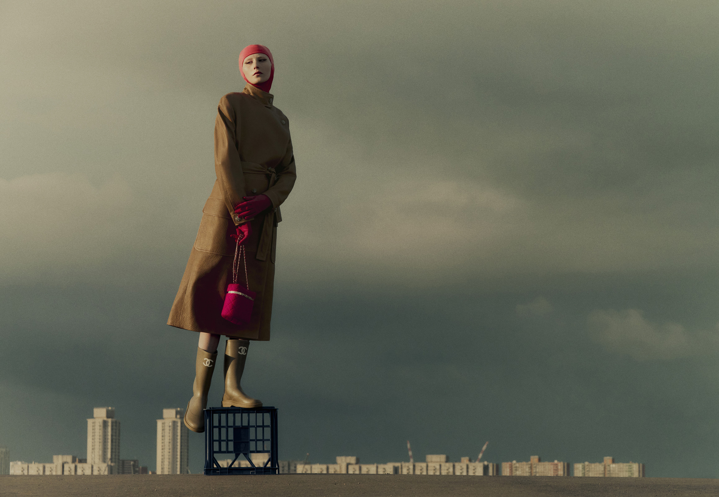 A fair-skinned female fashion model wearing a tan coat, pink gloves and beanie, standing on a crate holding a pink purse in an outdoor scene.