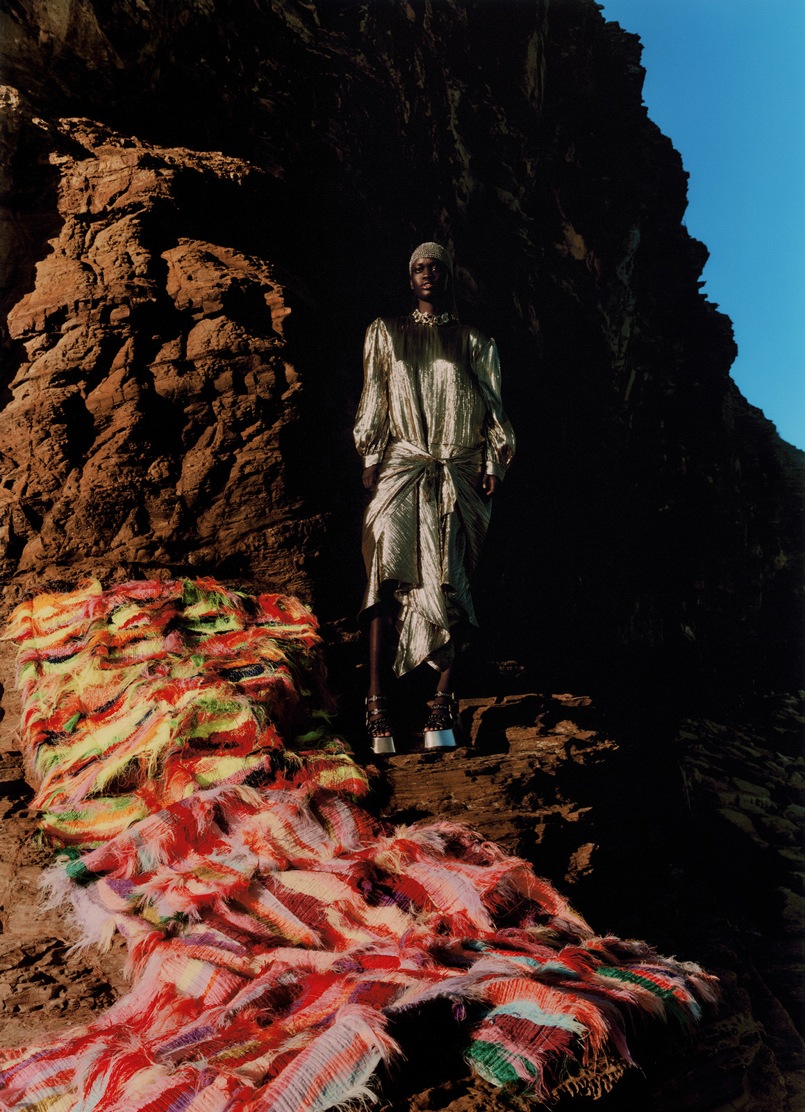 Fashion model poses on a rocky landscape wearing an elegant shiny dress and high heels, with a bright colorful rug in the foreground.