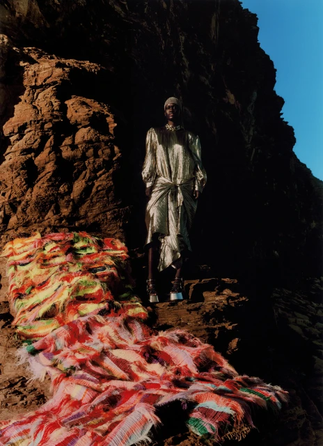 Fashion model poses on a rocky landscape wearing an elegant shiny dress and high heels, with a bright colorful rug in the foreground.
