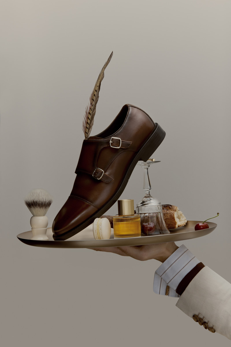 Studio product shot of brown leather monk-strap shoe on a silver tray with grooming accessories, liquor glass, bread, and cherries.