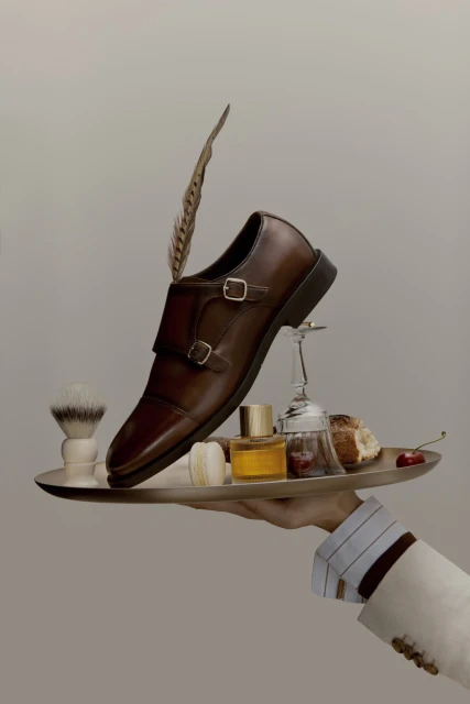 Studio product shot of brown leather monk-strap shoe on a silver tray with grooming accessories, liquor glass, bread, and cherries.