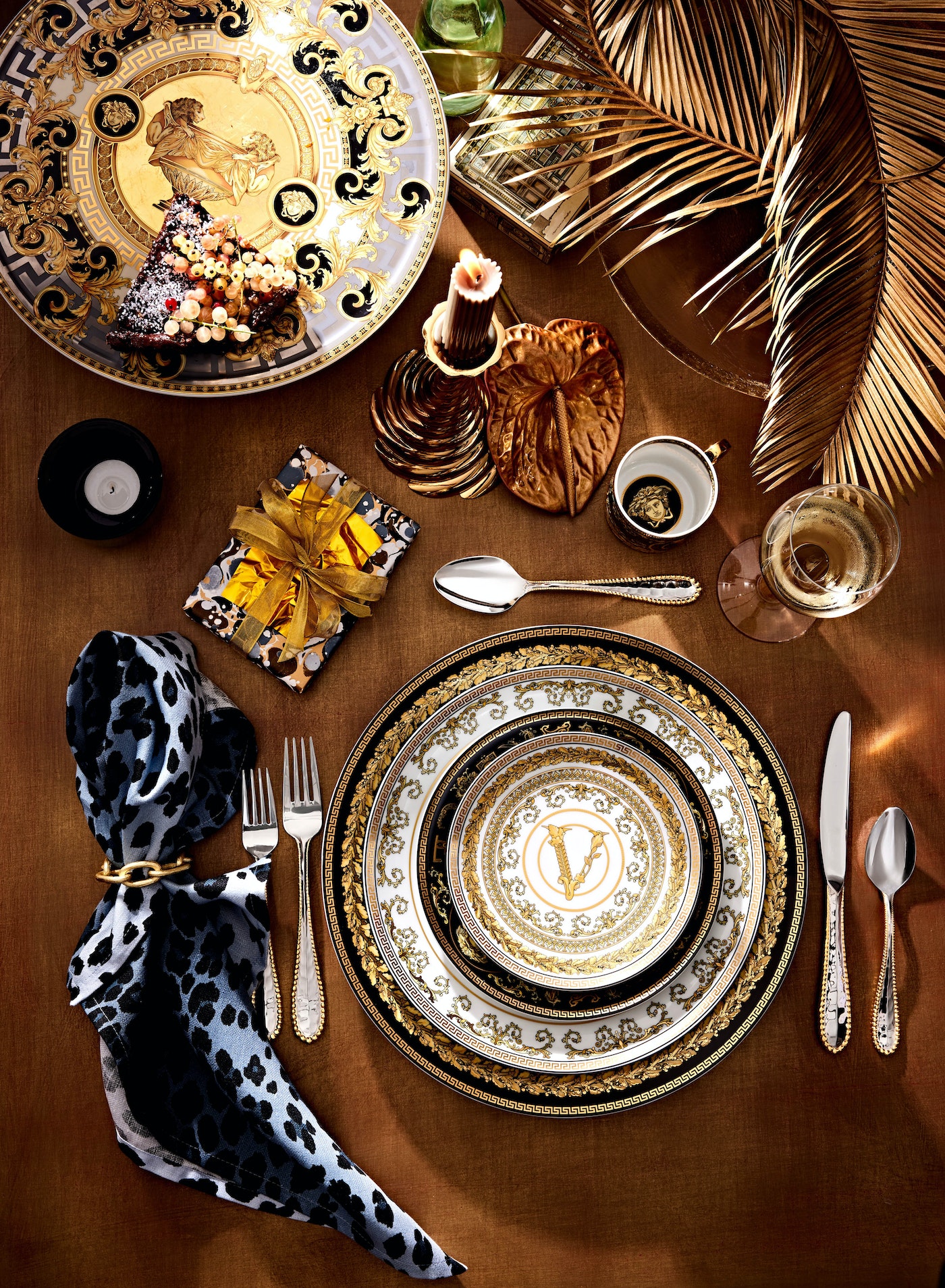 Overhead shot of a tabletop arrangement featuring Versace dinnerware, gifts, a candle, and golden decorative elements, opulent aesthetic.