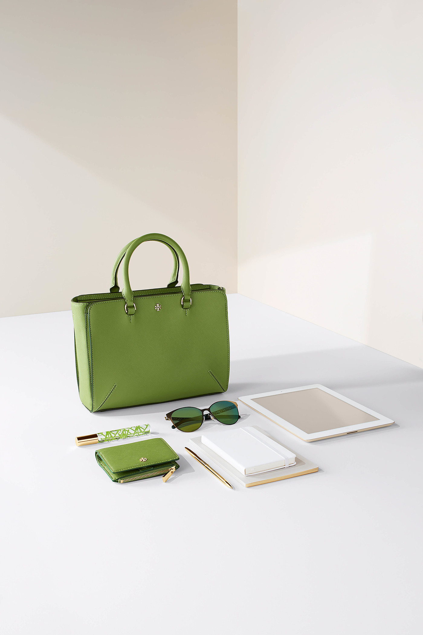 Studio shot of a lime-green leather handbag with matching wallet, sunglasses, a tablet, and stationary on a white surface against a neutral background.