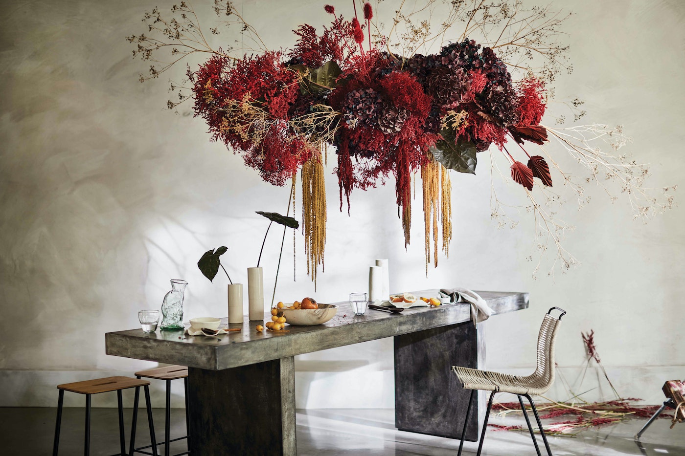 Floral centerpiece with hanging red and burgundy flowers above a concrete table set with dishes, fruit, and a modern chair in a bright room.