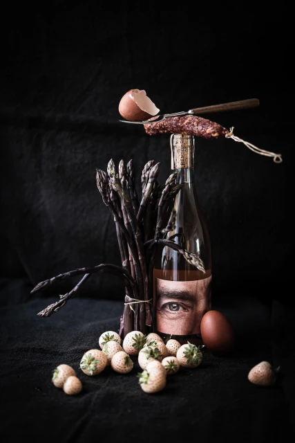 Still life with asparagus, strawberries, egg, and wine bottle featuring an eye on the label, all against a dark background.