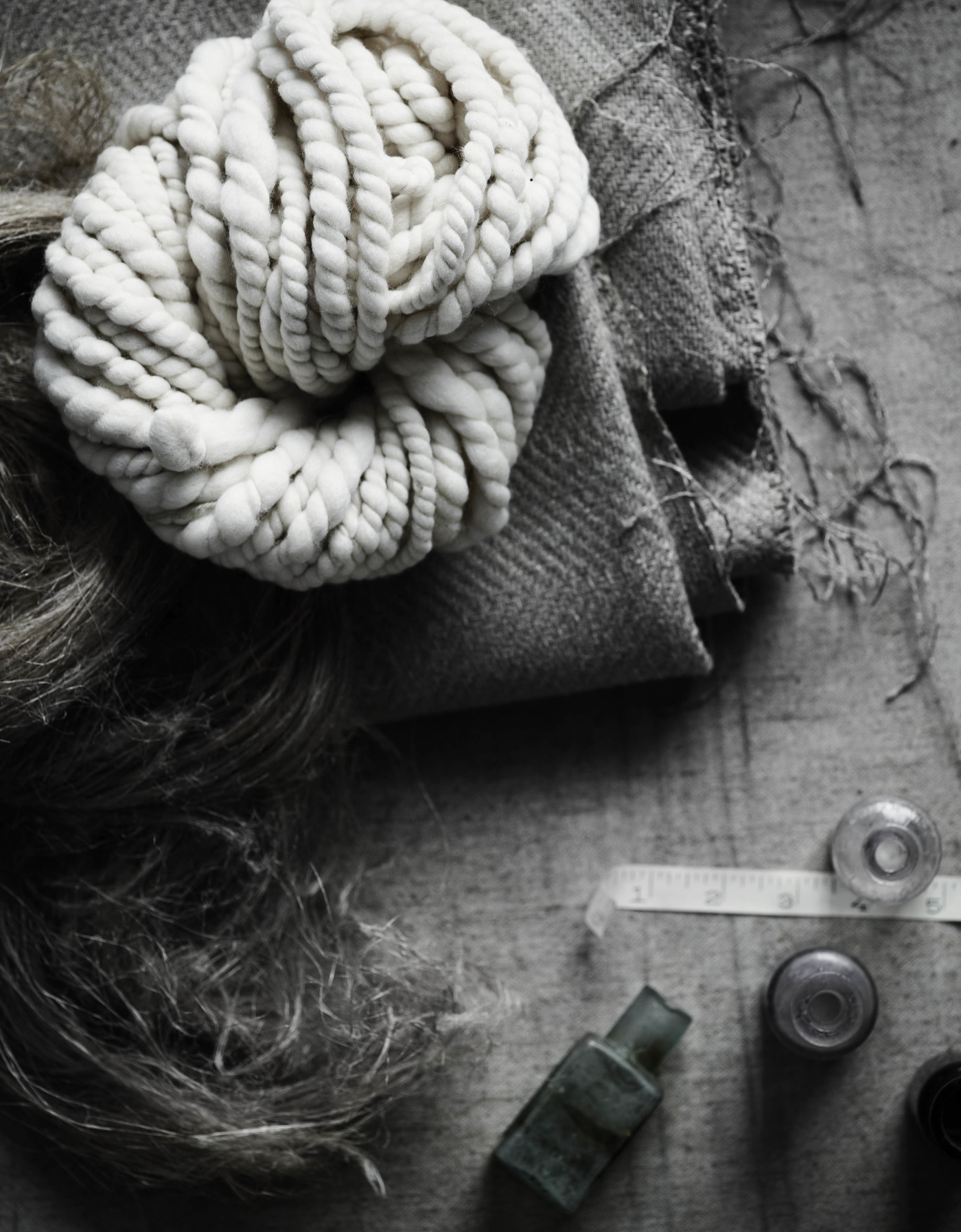Still life of woolen yarn, fabric swatch, and antique glass bottles on a wooden surface in muted monochrome tones, evoking a vintage aesthetic.