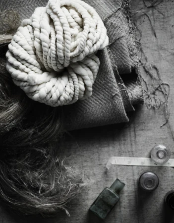 Still life of woolen yarn, fabric swatch, and antique glass bottles on a wooden surface in muted monochrome tones, evoking a vintage aesthetic.