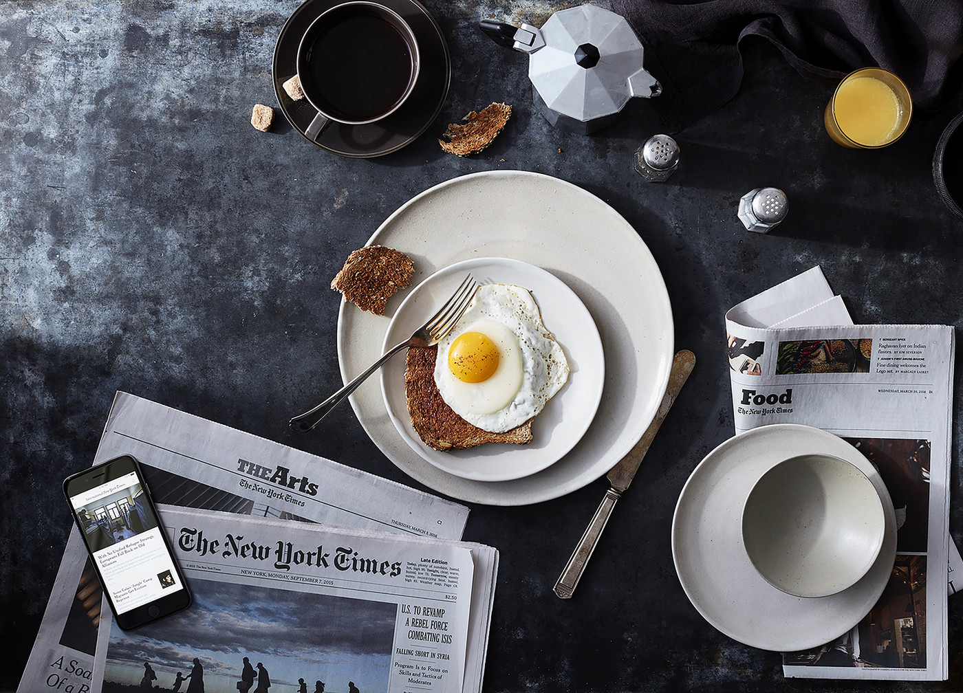 Overhead shot of a breakfast scene with coffee, toast, egg, juice, and newspapers on a dark textured surface, shot in horizontal format.