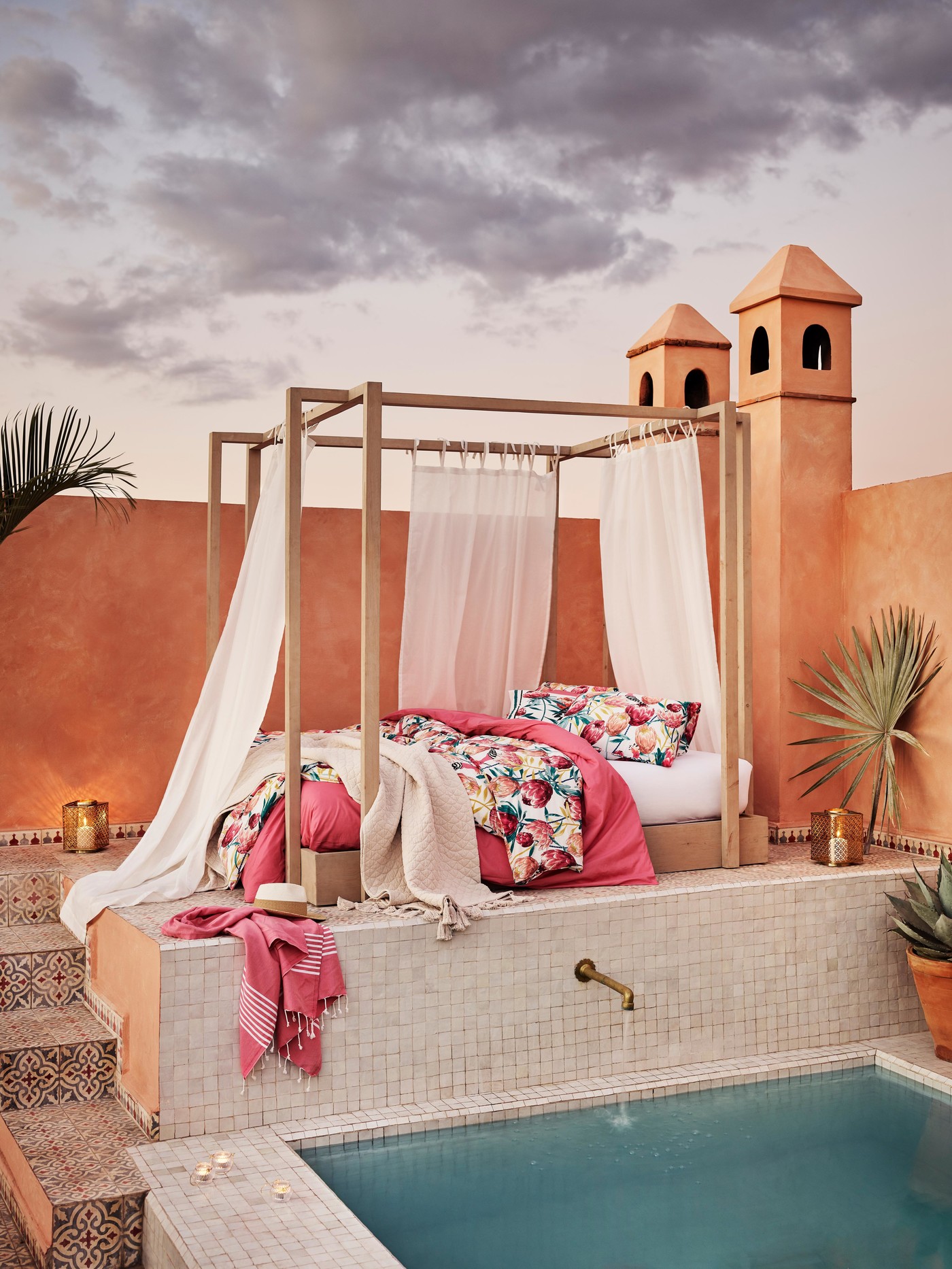A canopy bed on an outdoor terrace with a swimming pool, decorative towers, and an evening sky.