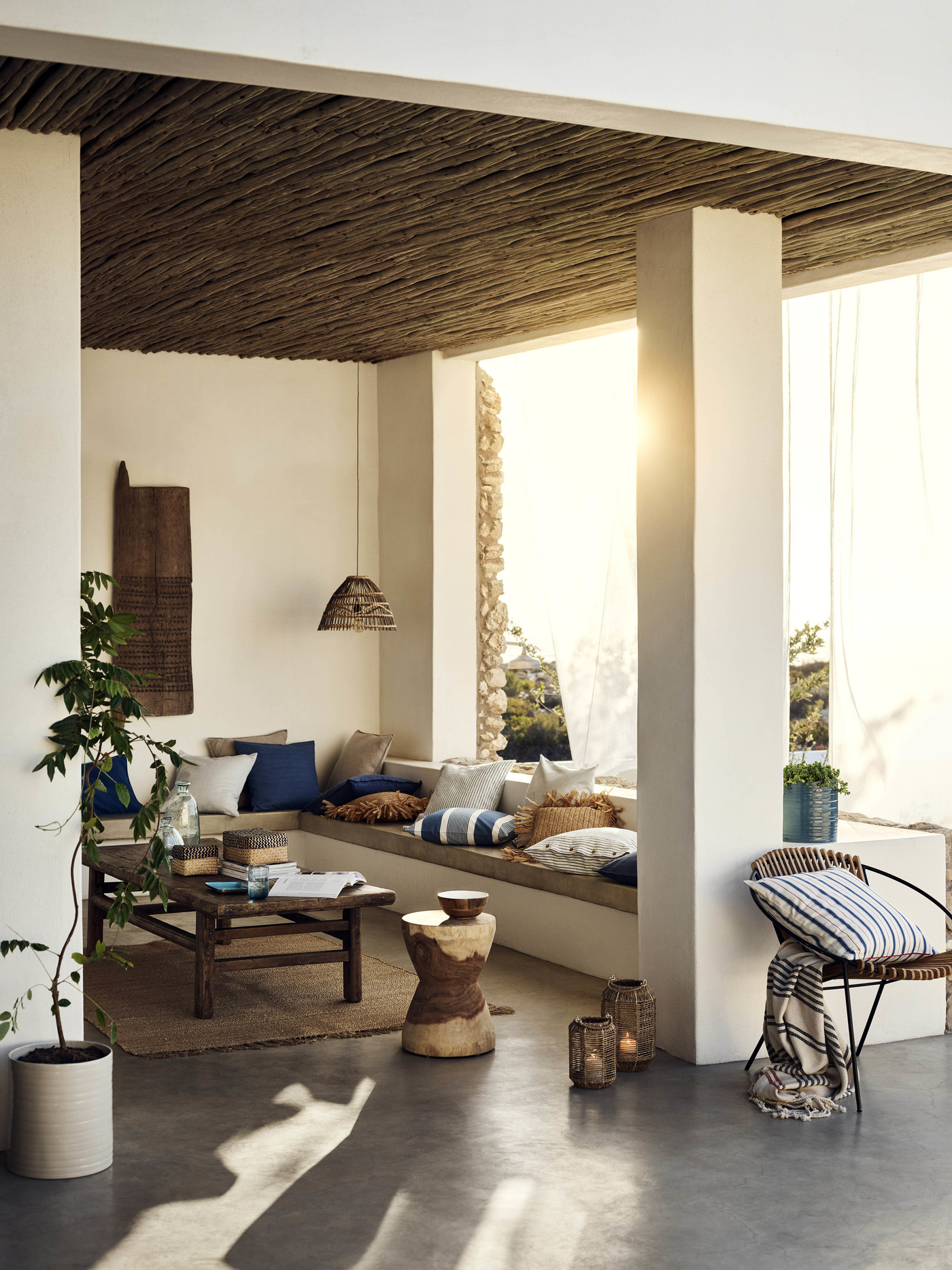 Sun-drenched terrace featuring built-in seating with pillows, rustic wood furniture, woven ceiling, and potted plants in a stylish outdoor living space.